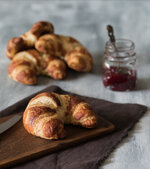 croissant auf holzbrett mit messer und marmelade im hintergrund auf hellem untergrund.jpg